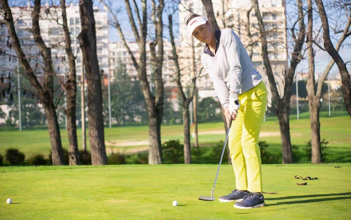 woman on a green with a putter wearing a light blue sweater, white visor, yellow pants
